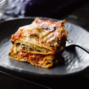 A close-up of a slice of vegan lasagna on a dark, round plate. The lasagna has visible layers of pasta, eggplant, tomato sauce, and melted cheese. A fork is placed on the plate next to the lasagna.